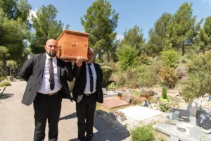 Deux porteurs funéraires transportant un cercueil en bois à l'épaule dans un cimetière.